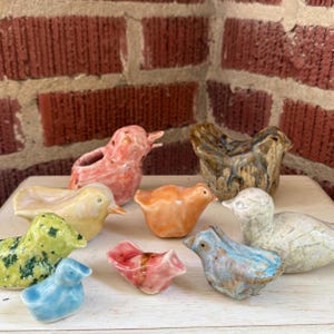 May include: A collection of eight small, glazed ceramic bird figurines in various colors including pink, yellow, green, orange, brown, blue, and white. Each bird features a smooth, glossy finish and distinct, rounded shapes, arranged together on a surface.