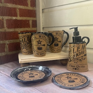 May include: A collection of tan ceramic kitchenware featuring black text and graphics. Items include two mugs, a soap dispenser, a small bowl, and a coaster, all labeled with Salem Witches Union or Salem Broom Co. and the year 1692.