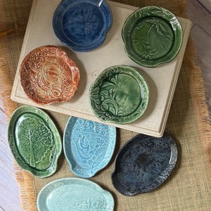 May include: A collection of eight ceramic dishes in various shapes and colors, including blue, green, and orange tones. Each dish features unique embossed patterns such as palm trees, floral motifs, birds, and foliage, with smooth glazed finishes.
