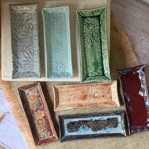 May include: Seven rectangular ceramic trays with varied glazes and textures. One features a rabbit, one has sunflowers, and others display intricate floral, swirling, or abstract patterns in shades of blue, green, red, and tan with glossy finishes.