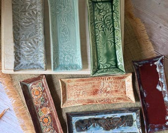 May include: Seven rectangular ceramic trays with varied glazes and textures. One features a rabbit, one has sunflowers, and others display intricate floral, swirling, or abstract patterns in shades of blue, green, red, and tan with glossy finishes.