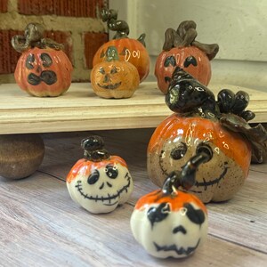 May include: A collection of small, glazed ceramic pumpkins featuring hand-painted jack-o'-lantern faces. The set includes various orange, speckled, and white pumpkins with dark stems, some with stitched mouth designs and others with carved-style expressions.