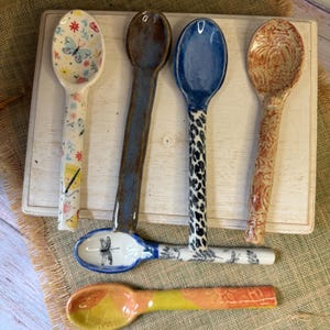 May include: Six ceramic spoons with distinct designs: one with butterflies and flowers, one solid dark brown, one with a blue bowl and leopard print handle, one with a textured orange finish, one with a dragonfly, and one with a yellow and orange pattern.