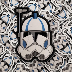May include: A collection of stickers featuring a white and blue helmet design. The central sticker is in focus, showcasing the helmet's details, while others are scattered around. The design includes blue stripes and a black visor.