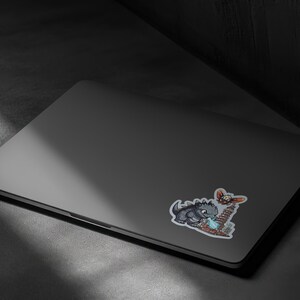 May include: A dark gray laptop featuring a sticker of a gray monster breathing blue fire at a tall building while a small winged creature flies above. The sticker has a white border and depicts a scene with a city skyline and a large creature.