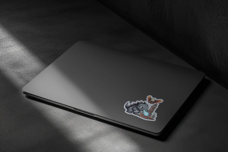 May include: A dark gray laptop featuring a sticker of a gray monster breathing blue fire at a tall building while a small winged creature flies above. The sticker has a white border and depicts a scene with a city skyline and a large creature.