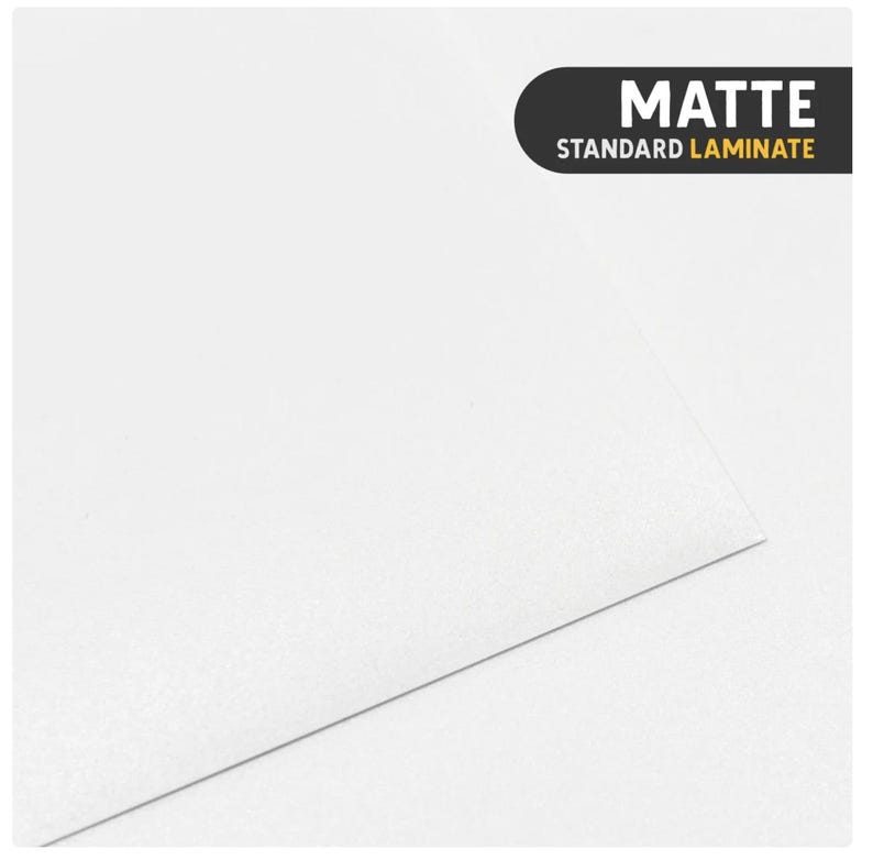 May include: A solid white sheet of material with a smooth, non-reflective surface. In the upper right corner, a black rectangular label with rounded corners features the white text MATTE and the yellow text STANDARD LAMINATE.