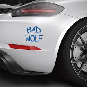 May include: A white car's rear section with a "BAD WOLF" decal in blue. The car has a black tire with a silver rim and orange brake calipers. A red reflector is visible.