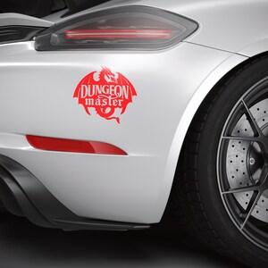 May include: A red "Dungeon Master" decal featuring a dragon design is affixed to the rear of a white car. The car's black tire and wheel are visible, along with a red taillight.