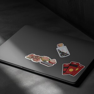 May include: A collection of stickers on a laptop. The stickers include a jar with a cork stopper, a cluster of food items, and two red and gold cards. The jar contains black and white confetti.