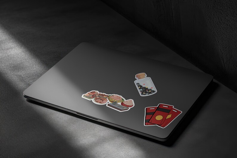 May include: A collection of stickers on a laptop. The stickers include a jar with a cork stopper, a cluster of food items, and two red and gold cards. The jar contains black and white confetti.