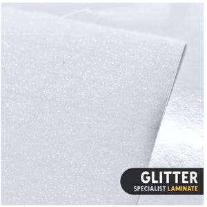 May include: A sheet of white glitter laminate with a fine, sparkling texture. The surface reflects light, creating a shimmering effect across the material. A black rectangular label in the corner displays the white text GLITTER and the yellow text SPECIALIST LAMINATE.