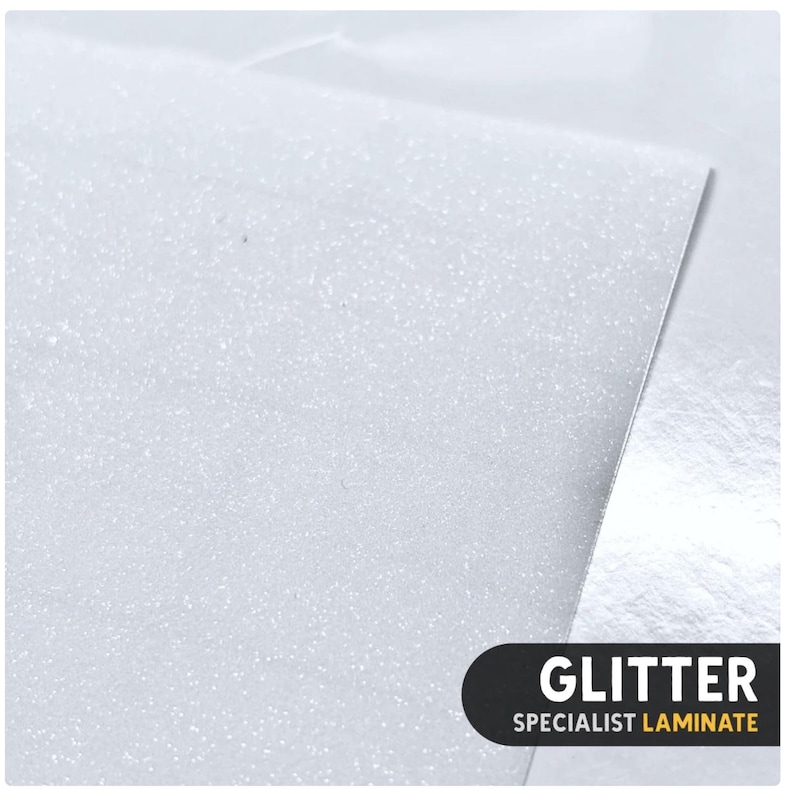 May include: A sheet of white glitter laminate with a fine, sparkling texture. The surface reflects light, creating a shimmering effect across the material. A black rectangular label in the corner displays the white text GLITTER and the yellow text SPECIALIST LAMINATE.