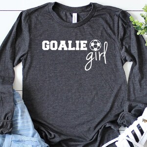 Op de afbeelding: Een donkergrijs shirt met lange mouwen met de tekst "GOALIE girl" en een witte voetbalafbeelding.