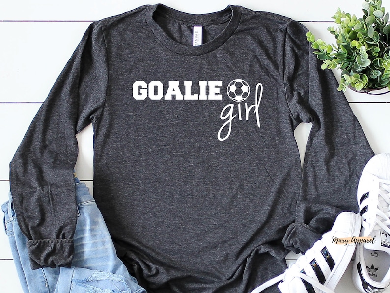 Op de afbeelding: Een donkergrijs shirt met lange mouwen met de tekst "GOALIE girl" en een witte voetbalafbeelding.