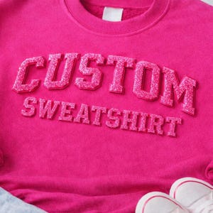 May include: A bright hot pink crewneck sweatshirt featuring the word CUSTOM in large, raised, glittery pink block letters, with the word SWEATSHIRT in smaller, matching glittery pink block letters underneath, all centered on the chest.