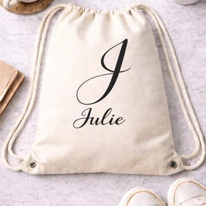 May include: A cream-colored drawstring bag with the name "Julie" and the letter "J" printed in black script. The bag has white drawstrings and metal eyelets. A pair of white sneakers is at the bottom.
