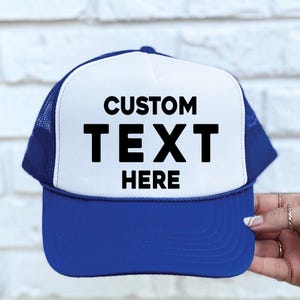 May include: A blue and white trucker hat with the words "CUSTOM TEXT HERE" in bold black letters. The hat has a white front panel, a blue brim, and a blue mesh back. A hand is holding the hat.