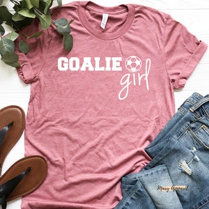 Op de afbeelding: Een roze T-shirt met de tekst "Goalie girl" in wit met een witte voetbalafbeelding. Het shirt ligt op een wit oppervlak met een paar bruine sandalen en een blauwe spijkerbroek.