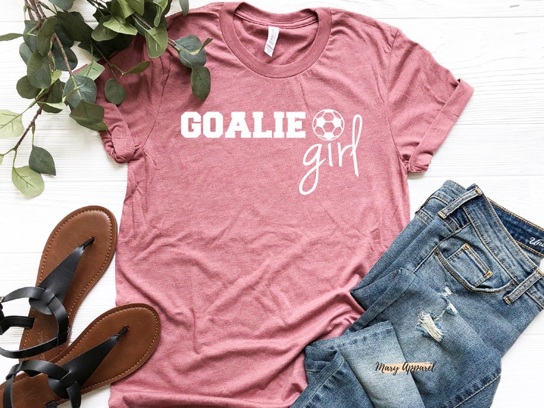 Op de afbeelding: Een roze T-shirt met de tekst "Goalie girl" in wit met een witte voetbalafbeelding. Het shirt ligt op een wit oppervlak met een paar bruine sandalen en een blauwe spijkerbroek.