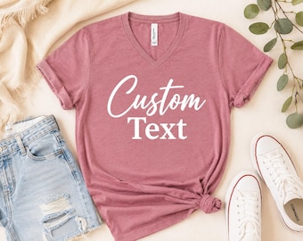 May include: A mauve heathered V-neck t-shirt featuring the white text Custom Text printed in a combination of script and serif fonts on the center chest. The shirt has short sleeves and a standard hemline, presented as a flat lay garment.