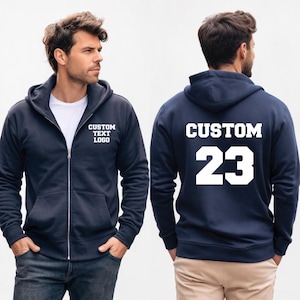 May include: Navy blue zip-up hoodie with white text that reads "CUSTOM TEXT LOGO" on the front and "CUSTOM 23" on the back. The hoodie has a hood and pockets. The model is wearing blue jeans.