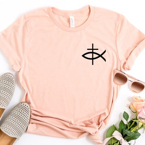 May include: A peach-colored t-shirt with a black Christian fish symbol on the front. The shirt is laid flat on a white surface with a pair of beige shoes, sunglasses, and a bouquet of pink roses.