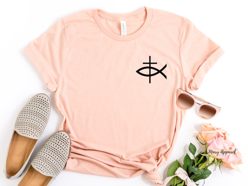 May include: A peach-colored t-shirt with a black Christian fish symbol on the front. The shirt is laid flat on a white surface with a pair of beige shoes, sunglasses, and a bouquet of pink roses.