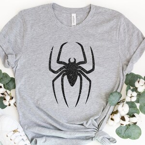 K&ouml;nnte beinhalten: Ein graues T-Shirt mit einem schwarzen Glitzer-Spinnen-Design.