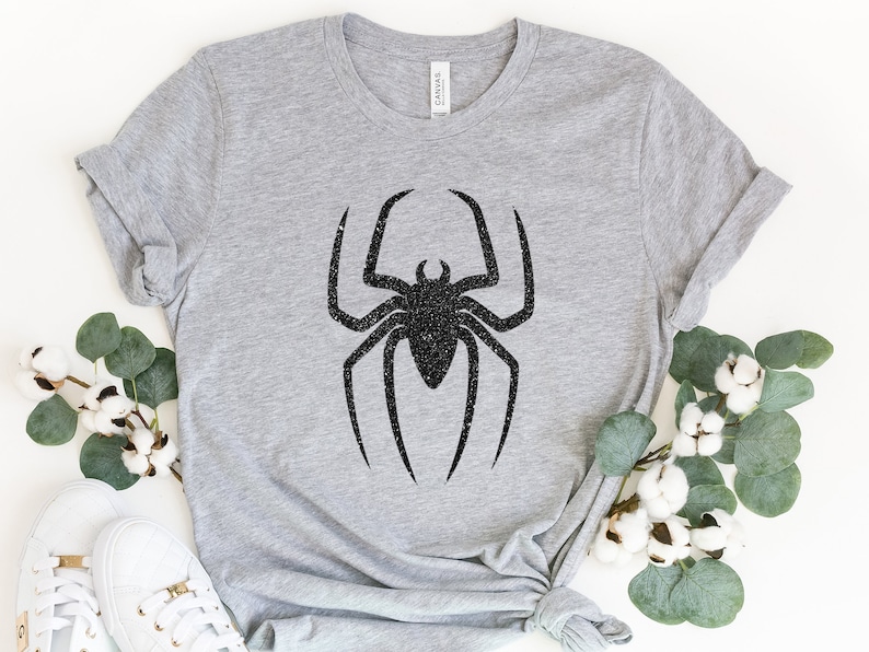 K&ouml;nnte beinhalten: Ein graues T-Shirt mit einem schwarzen Glitzer-Spinnen-Design.