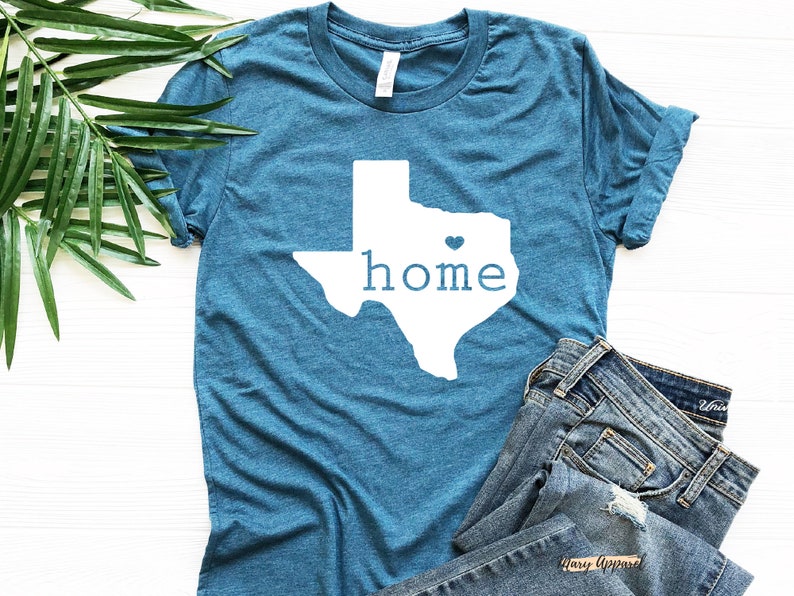 May include: A teal blue t-shirt with a white outline of the state of Texas and the word "home" inside. A small white heart is in the center of the state.