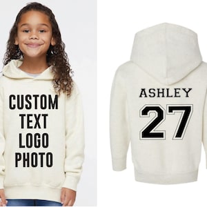 A young girl wearing a white hoodie with the text "CUSTOM TEXT LOGO PHOTO" printed on it, and the name "ASHLEY 27" printed on the back.
