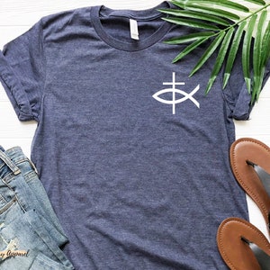May include: A navy blue t-shirt with a white fish and cross graphic. The shirt is laid flat on a white wooden surface with a pair of brown sandals and a pair of blue jeans.