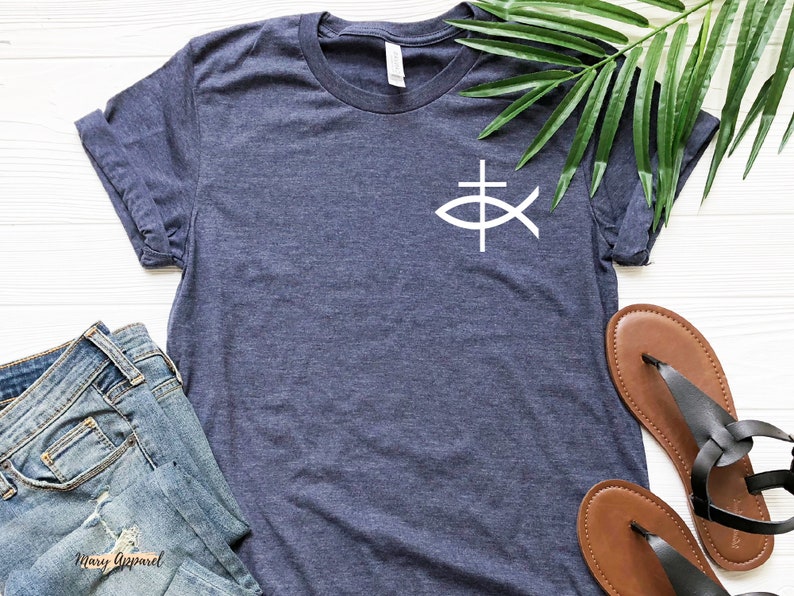 May include: A navy blue t-shirt with a white fish and cross graphic. The shirt is laid flat on a white wooden surface with a pair of brown sandals and a pair of blue jeans.