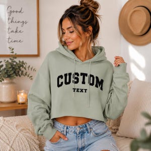 May include: Sage green cropped hoodie with the word "CUSTOM" arched across the chest in bold black letters, with "TEXT" below. The hoodie has a drawstring hood and long sleeves. The model is wearing blue jeans.