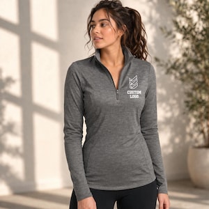 May include: A gray long-sleeve athletic top with a quarter-zip neckline. The top features a white logo and the text "CUSTOM LOGO" on the chest. The model is wearing black athletic pants. The background includes a plant.