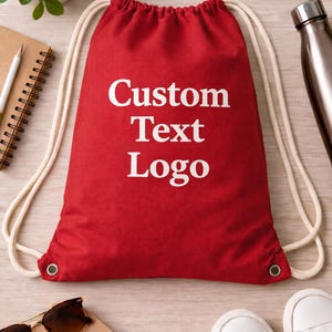 May include: Red drawstring bag with white text reading "Custom Text Logo." The bag has white drawstrings and metal eyelets. Other items include sunglasses, a notebook, a pencil, white sneakers, and a water bottle.