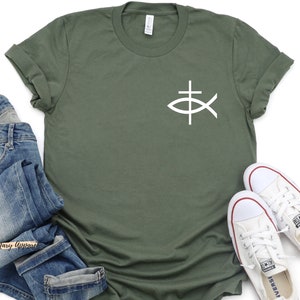 May include: A green t-shirt with a white fish symbol with a cross on top. The shirt is folded and laid on a white surface with a pair of white sneakers and blue jeans.