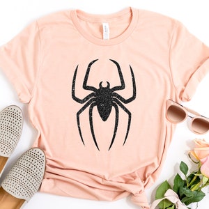 K&ouml;nnte beinhalten: Ein hellrosa T-Shirt mit einem schwarzen Glitzer-Spinnen-Motiv. Das Shirt liegt flach auf einer wei&szlig;en Oberfl&auml;che mit einem Paar wei&szlig;en Schuhen, einer Sonnenbrille und rosa Rosen.