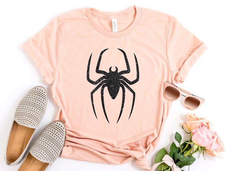 K&ouml;nnte beinhalten: Ein hellrosa T-Shirt mit einem schwarzen Glitzer-Spinnen-Motiv. Das Shirt liegt flach auf einer wei&szlig;en Oberfl&auml;che mit einem Paar wei&szlig;en Schuhen, einer Sonnenbrille und rosa Rosen.
