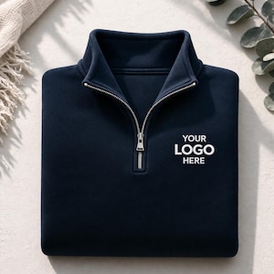 May include: A navy blue quarter-zip sweatshirt with a silver zip and a mock neck collar. The text "YOUR LOGO HERE" is printed in white on the front. The sweatshirt is folded neatly on a light-coloured surface.
