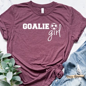 Op de afbeelding: Een maroon t-shirt met witte tekst die "Goalie girl" zegt en een witte voetbalafbeelding.