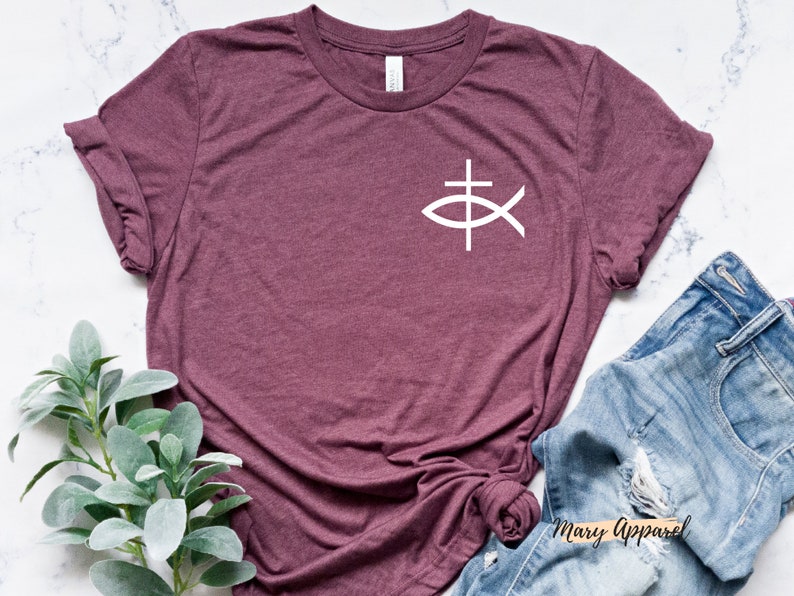 May include: A maroon t-shirt with a white fish symbol with a cross inside. The shirt is laying on a white marble surface with a pair of blue jeans and a green plant.
