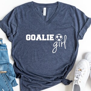 Op de afbeelding: Een marineblauw V-hals T-shirt met de tekst "GOALIE girl" in wit, met een witte voetbalafbeelding.