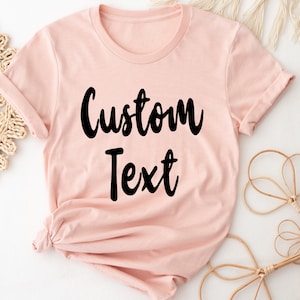 May include: A light pink t-shirt with the text "Custom Text" printed in black.