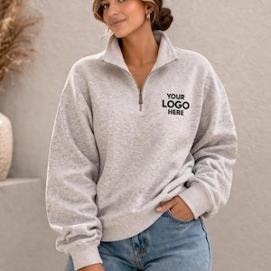 May include: A light gray sweatshirt with a quarter-zip neckline and the text "YOUR LOGO HERE" in black. The sweatshirt has long sleeves and a relaxed fit. The model is wearing blue jeans.
