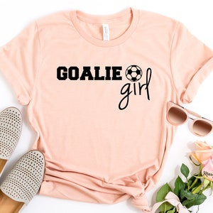 Op de afbeelding: Een perzikkleurig T-shirt met de tekst "GOALIE girl" in zwarte letters en een zwart-witte voetbalafbeelding. Het shirt ligt plat op een wit oppervlak met een paar beige schoenen, een zonnebril en roze bloemen.