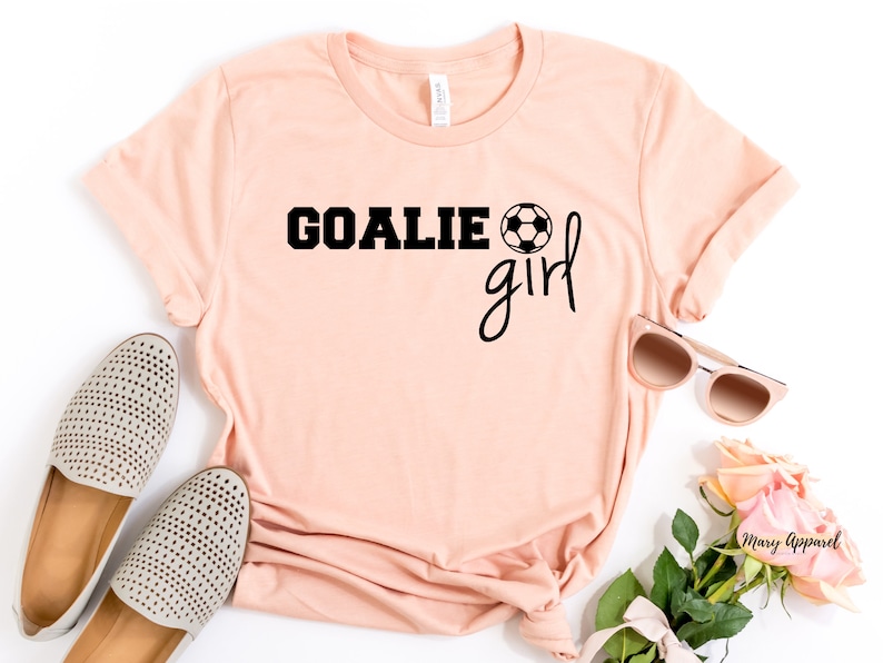 Op de afbeelding: Een perzikkleurig T-shirt met de tekst "GOALIE girl" in zwarte letters en een zwart-witte voetbalafbeelding. Het shirt ligt plat op een wit oppervlak met een paar beige schoenen, een zonnebril en roze bloemen.