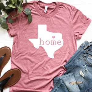 May include: A pink t-shirt with a white outline of the state of Texas and the word "home" inside. A small pink heart is in the center of the state outline. The shirt is laid flat on a white surface with a pair of brown sandals and blue jeans.