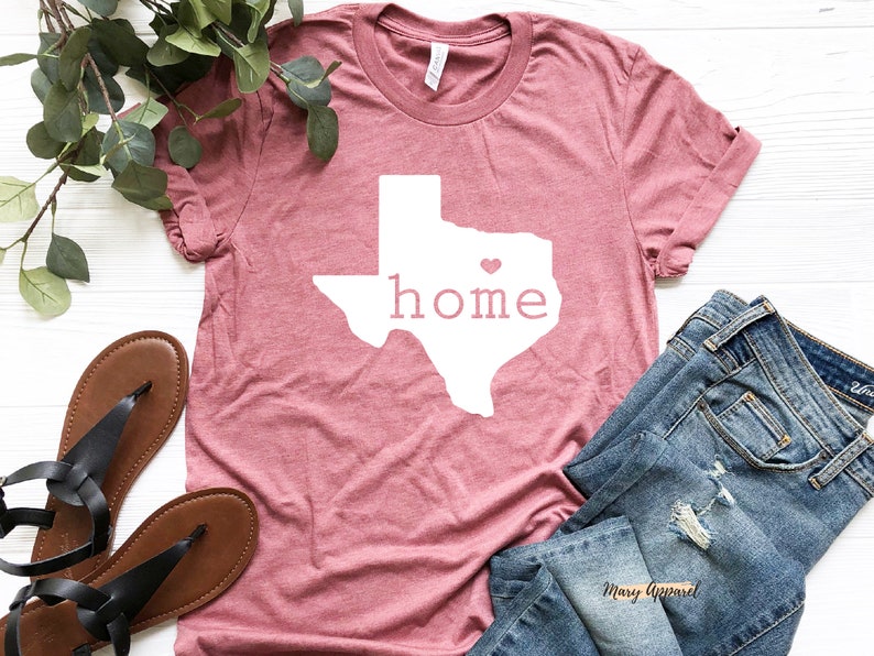 May include: A pink t-shirt with a white outline of the state of Texas and the word "home" inside. A small pink heart is in the center of the state outline. The shirt is laid flat on a white surface with a pair of brown sandals and blue jeans.